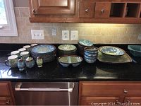 Full view of dishware set laid out on black countertop including plates, bowls, mugs, salt and pepper shakers, sugar bowl, serving bowls, and tray.