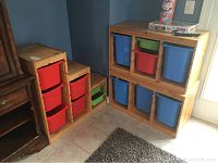 Three wooden storage units in natural finish with colorful plastic bins (red, green, blue) placed on tiled floor against blue wall.