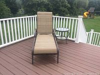 Outdoor metal and fabric chaise lounge with beige woven fabric seating and metal frame, facing front on an outdoor deck near white railing.
