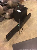 Front view of black soundbar and wireless subwoofer on concrete floor, showing both components and power cable.