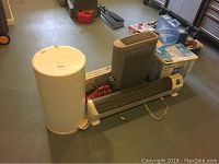 Photo showing white cylindrical Holmes Puremist humidifier, white trash bin, horizontal baseboard style heater, Honeywell tall vertical heater, and boxed Shop Light on grey floor.