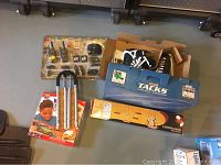 Lot overview showing all items together on floor including hockey skates in box, 2-way radios in original packaging, wooden fishing rod rack box, Balance Buddy training aid with packaging, and wooden car set box.