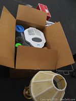 Top view inside open box showing white Oster deep fryer, glass lid, blue and green plastic lids or covers and parts of a decorative lamp base with floral shade.