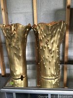 Front view of both gold-painted wooden vases on mirrored surface against concrete block wall