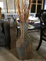 Front and angled view of tall metal floor vase with geometric circular and square designs and dried decorative stems inside.