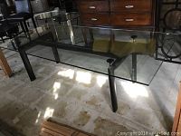 Front side view of the glass dining table showing the clear glass top, the steel legs, and the floor beneath.
