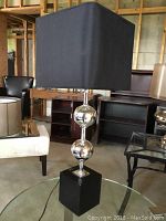 Full view of chrome table lamp on glass table showing chrome spherical accents and black rectangular base with black square shade.
