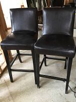 Front view of two dark brown leather upholstered bar stools with padded seats and backs, wooden legs showing some wear and minor scratches.