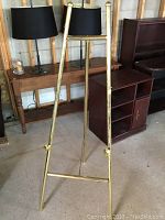 Full view of brass tone easel approx 60 inches tall, standing against a wall beside furniture and table lamps.