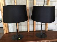 Pair of black metal table lamps with black drum shades seen together on a wooden surface against a wood and cement background.