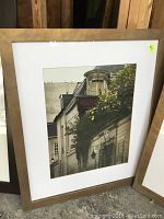 Framed artwork depicting an architectural scene with buildings and foliage, in a wooden frame with some visible wear.
