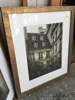 Photo of framed art print depicting an old building facade with architectural details and multiple windows. The frame is gold-toned wood with some visible wear marks and scratches.