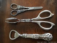 Photo showing three sterling silver-handled serving implements including scissors, tongs, and a bottle opener on a wooden surface.