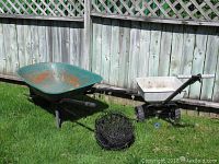 Overall view of all items: green rusted metal wheelbarrow, white tow-behind spreader, and pile of black wire hanging pots on grass in front of wooden fence.