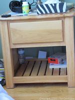 Photo of one pine night stand showing single drawer front and slatted bottom shelf with water bottle and box on shelf. Wear is visible on top.