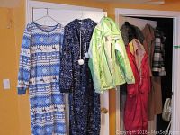 Seven pieces of ladies clothing hanging on a door including onesies, jackets, and coats in various colors and patterns.