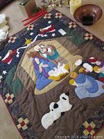 Close-up photo of the Nativity scene quilt with fabric appliqué art depicting Mary, Joseph, Baby Jesus, Wise Men, and animals on carpeted floor next to candles and other decor.