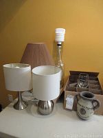 Photo of two table lamps with white shades, beige ceramic lamp with a crumpled beige shade, Schenley bottle with white shot pump, box of 12 Strongbow shot glasses, and decorative jug.