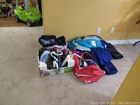 Overall view of mixed clothing lot including folded garments, a red duffel bag, and box with purses and shoes arranged on carpeted floor.