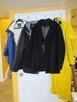 Four men's jackets and yellow rain gear pants hanging on a rack. Items include a black pea coat, two outdoor jackets, and yellow rain pants.