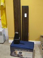 Wood grain finish single bed frame panels and black bed elevator feet on the floor against a yellow wall, folding bed wedge bag blue in color.