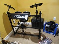 Front view of Alesis drum kit with Squier amp at rear and box of cables/pedals on floor