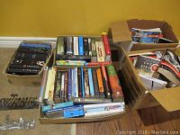 Three cardboard boxes filled with mixed hardcover and paperback books, visibly aged, and two small metal signs on floor reading 'Swimming' and 'Hockey'.