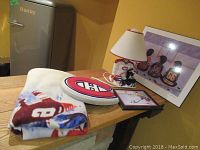 Photo showing hockey blanket with Canadiens logo, white Canadiens logo plaque, framed print of kids in hockey jerseys, and collectible hockey item sets including helmet and skates next to lamp