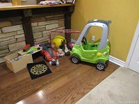 Wide angle shot showing all items in the lot arranged on a wooden floor against a stone wall and yellow wall, includes ride-on cars, net, balls, dartboard, and stuffed animals.