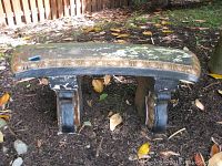 Full view of the curved outdoor garden bench positioned on mulch ground with some leaf litter, showing the decorative edging and two pedestal legs.