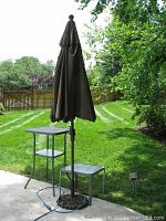 Outdoor set on patio showing closed large brown fabric umbrella, tall rectangular table and smaller square table on a concrete patio with lawn and trees in the background.