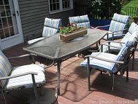 Full patio set arranged outside on wooden deck, showing six chairs around rectangular glass topped table with metal frame and umbrella stand base.