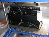 Adcom amplifier and Optimus 5-disc CD changer stacked on metal shelving, with power cords visible. Signs of wear and storage conditions.