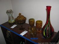Overview of six amber ware decorative glass items including vases and goblets arranged on a shelf.