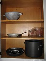 View of kitchen shelf with colander, frying pan, black pots with lids, and Pyrex measuring cups