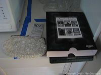 Amazon Kindle Paperwhite e-reader box and white beaded purse side by side on countertop.