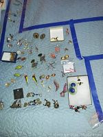 Wide angle photo showing assortment of costume earrings laid out on a blue quilted surface, various colors, shapes, and sizes visible