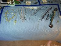 Assorted costume jewelry including multiple necklaces of various styles laid out on quilted blue bedding. Necklaces show a range of chain types and charm pendants.