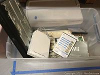 Photo showing white Nintendo Wii console inside a plastic container along with cables and manuals.