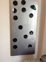 Metal magnetic board with multiple round black magnets and one green clip magnet attached on a wall.