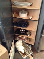 Shelf with white ceramic casserole dish with lid (chip on lid), white platters on upper shelf, clear Pyrex baking dishes, stainless steel and metal mixing bowls, and assorted items stored below including linens and placemats.