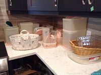 Wide shot showing large set of translucent Tupperware containers with pastel lids and a white ceramic decorative basket with floral decoration on a kitchen counter