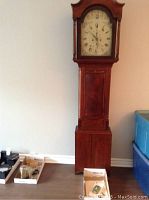 Full view of mahogany-veneered longcase clock standing against wall with accessories beside it