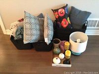 Photo showing five black woven baskets filled with various throw pillows and blankets, arranged beside a small lamp and candles on the floor.