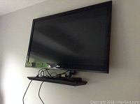 Front view of black LG flat screen TV mounted on wall with cables hanging and small shelf underneath.
