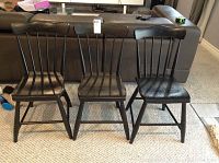 Full view of the three black painted wooden chairs with spindle backs arranged side by side on carpeted floor with sofa in the background.