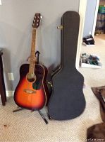 Overall view of the Tradition sunburst acoustic guitar on the Ultra stand with the black hard shell case next to it