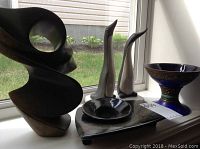 Modern abstract sculpture with smooth curves and hollow center placed on a window sill with penguin figurines, flower holder, and blue dish nearby.