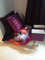 Photo showing queen size comforter with matching pillow shams and two decorative throw pillows with textured fabric in purple and black, placed on carpeted floor next to black curtain.