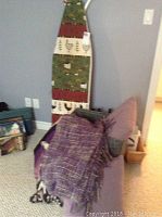Photo showing a decorative ironing board with painted farm scenes including a chicken and corn, along with stacked pillows and a woven blanket draped on a chair.